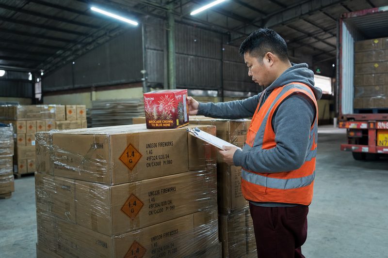 Private-label and OEM fireworks — a warehouse worker in a high-visibility vest checking a branded consumer firework box against a clipboard, beside shrink-wrapped pallets of UN0336 1.4G export cartons